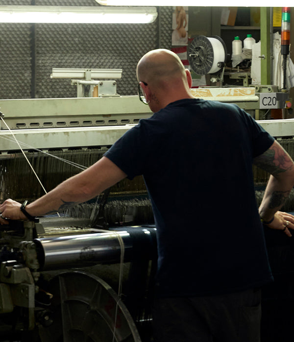 A worker viewed from behind, leaning over an industrial loom and guiding fabric through the machine in a textile factory.